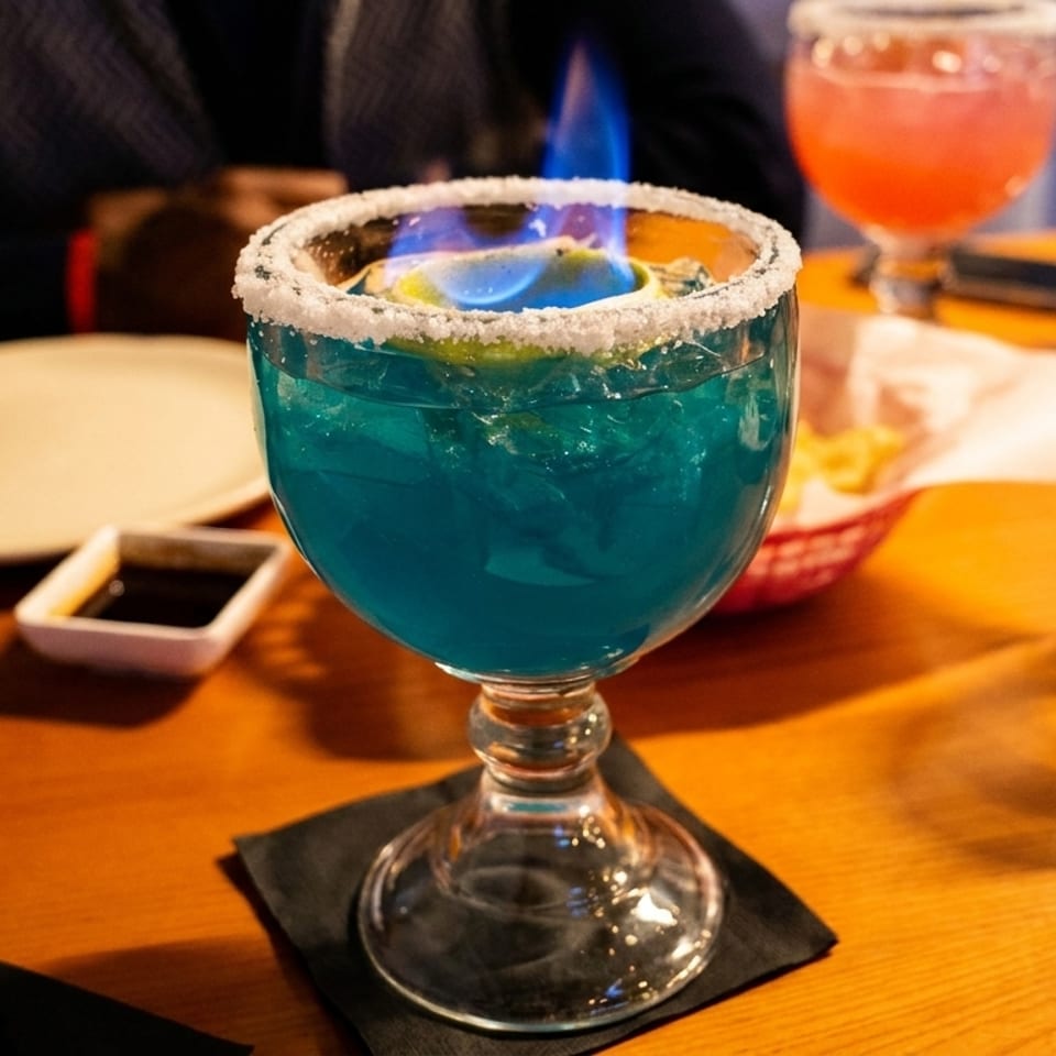 A blue flaming cocktail with a salted rim and lime sits on a wooden table.