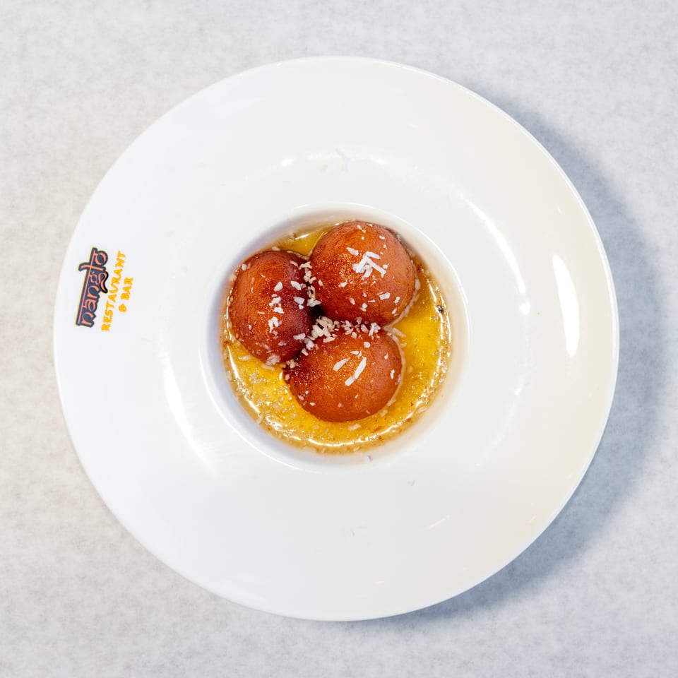 Three syrup-soaked brown dessert balls topped with shredded coconut, served in a white Rajdhani Restaurant & Bar dish.