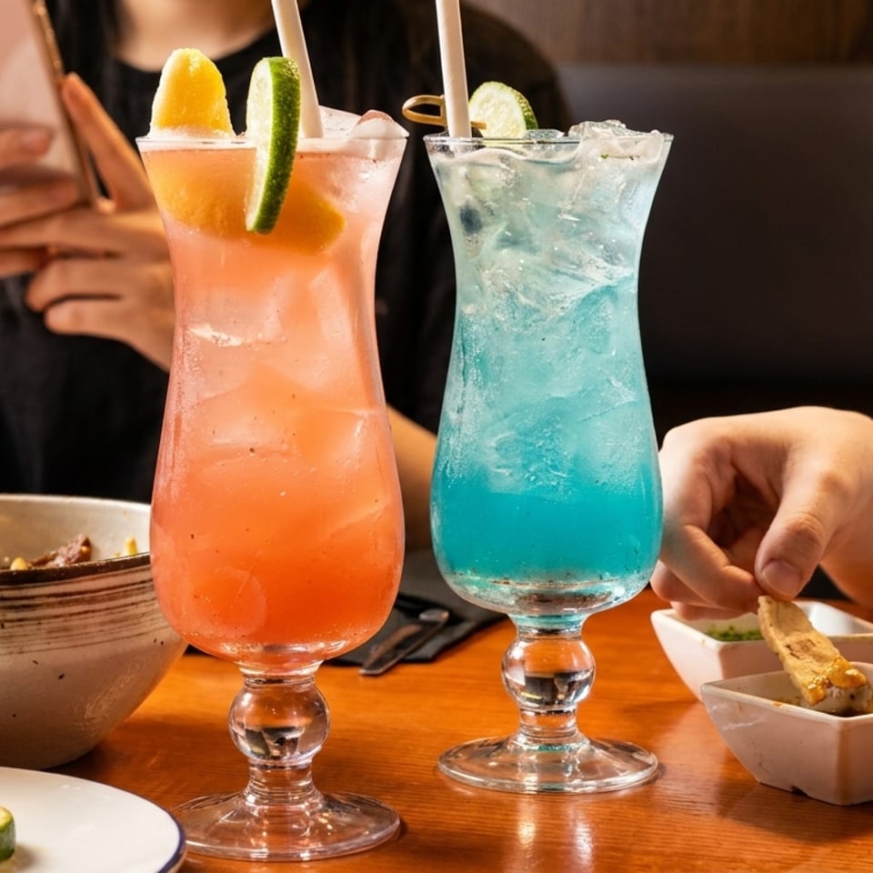 Two people at a table with colorful cocktails, noodles, and a person dipping food.