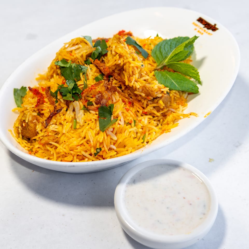 Spiced chicken biryani garnished with mint, cilantro, and crispy onions, served with a bowl of creamy raita.