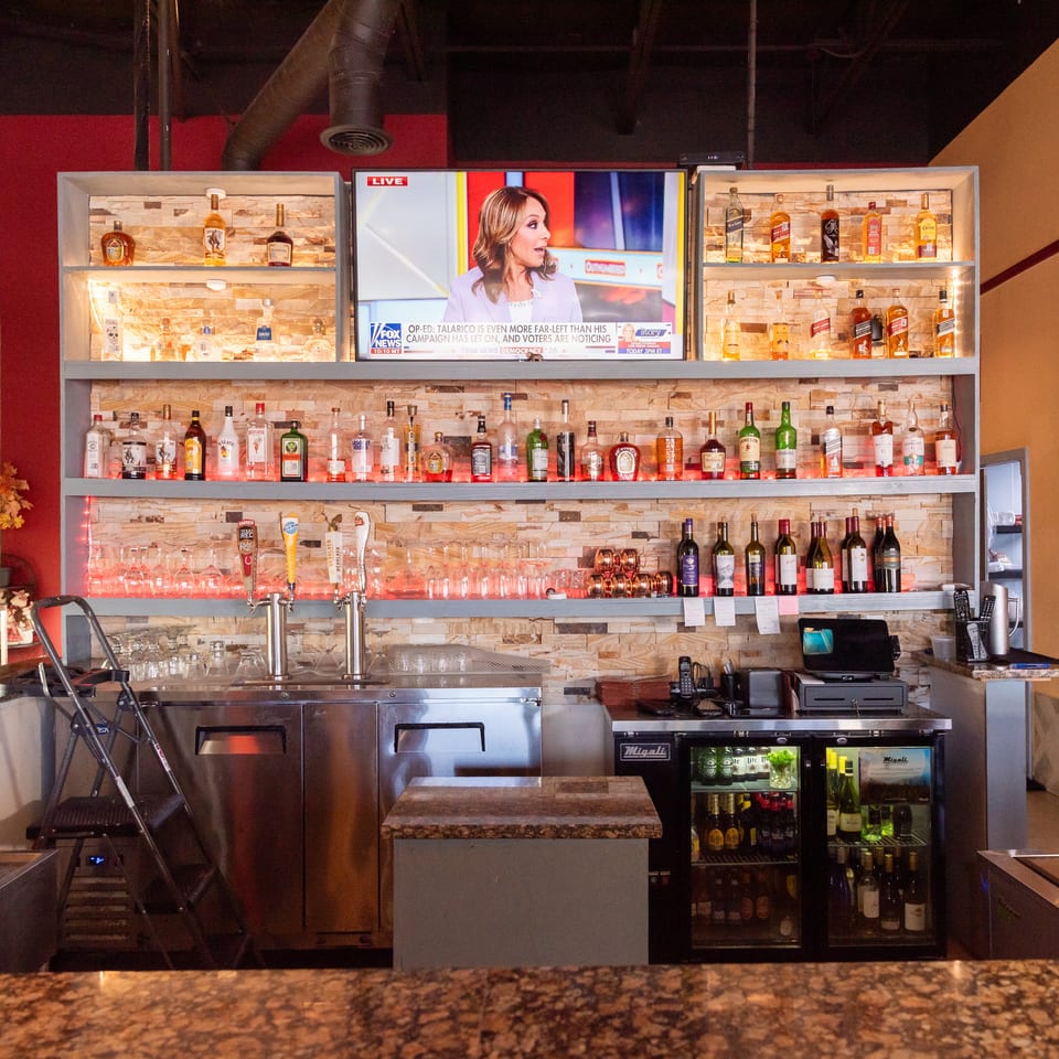Restaurant bar with illuminated shelves of liquor bottles, wine, beer taps, glassware, television, and refrigerators.