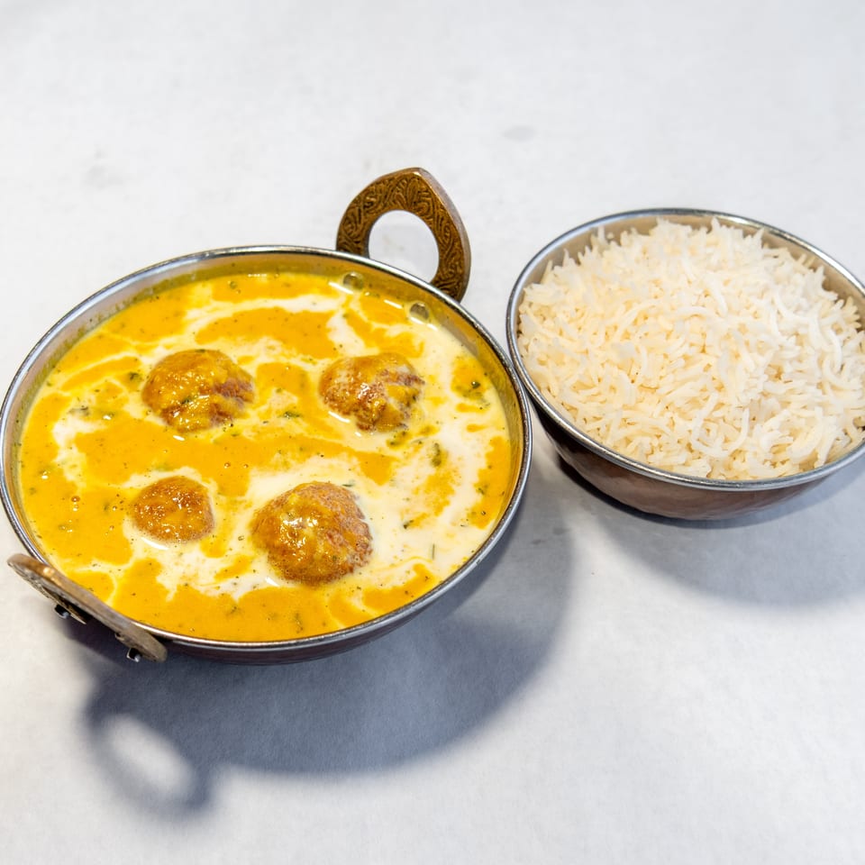 Indian kofta curry in a decorative serving pan with cream sauce, accompanied by a bowl of basmati rice.