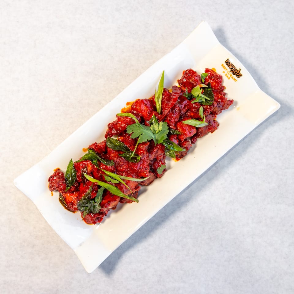 Red-glazed fried chicken pieces garnished with cilantro, curry leaves, and green onions on a white rectangular plate.