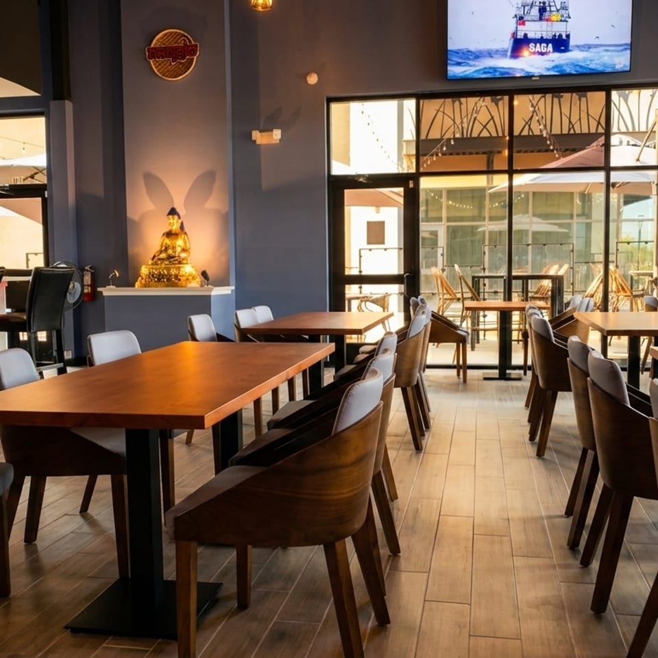 Modern restaurant interior with wooden tables, gray chairs, a bar, and a golden Buddha statue.