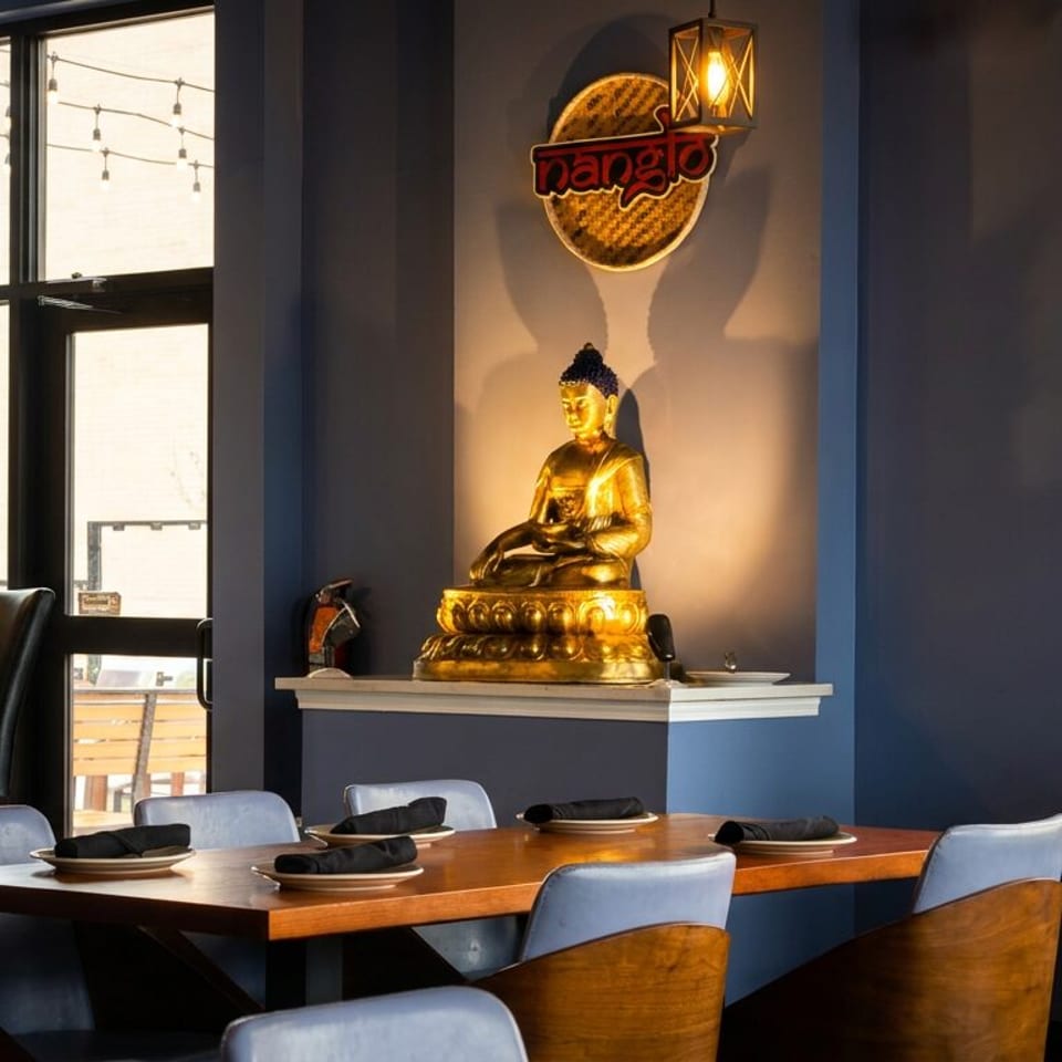 Restaurant interior with a golden Buddha statue, Nanglo sign, wooden tables, and blue chairs.