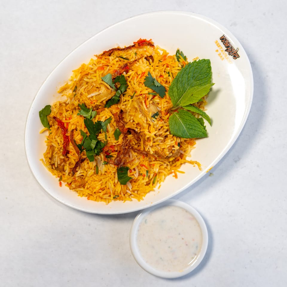Biryani with spiced rice, chicken, fried onions, cilantro, and mint, served with a small bowl of raita.