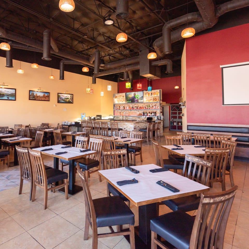 Spacious restaurant dining room with set tables, wooden chairs, warm pendant lighting, a stocked bar, and projection screen.